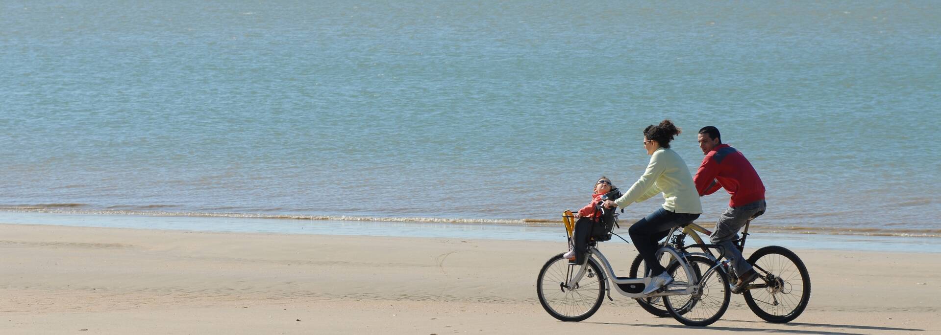 Deux personne et leur enfant qui font une balade à vélo sur le sable au bord de l'océan